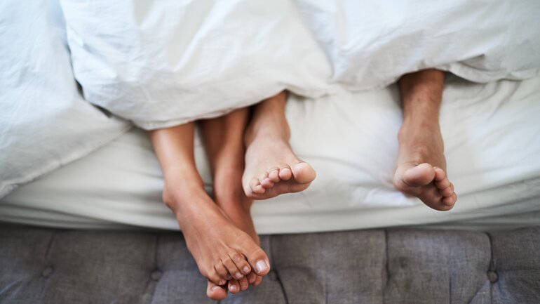 Zwei Personen liegen im Bett, nur die Füße schauen unter der Bettdecke hervor
