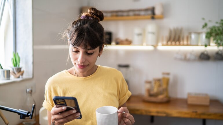 Junge Frau mit Tattoos am Arm und messy bun steht in der Küche und schaut auf ihr Smartphone