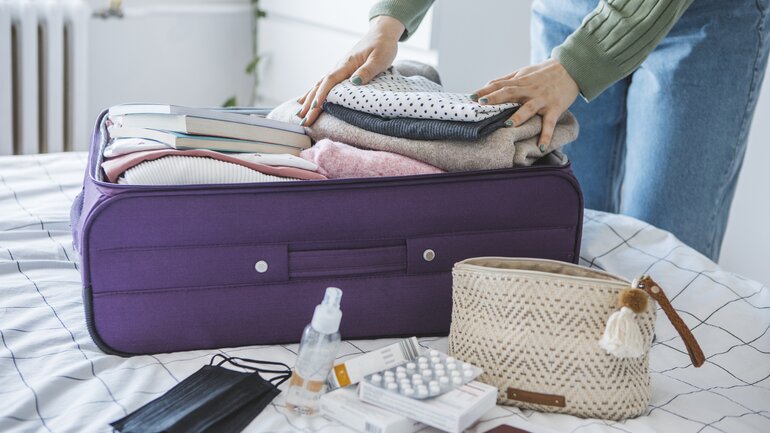 Frau packt ihren Koffer für eine Reise, im Vordergrund sind auch medizinische Masken und Medikamente zu sehen