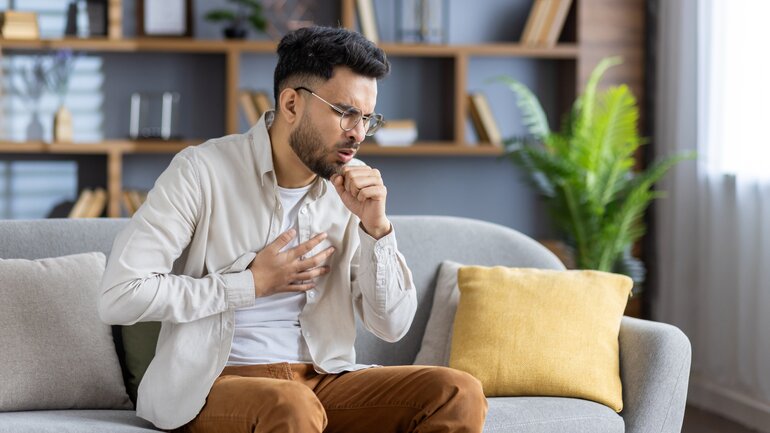 Junger Mann mit Brille sitzt daheim auf dem Sofa und muss stark husten, er hält sich eine Hand vor Schmerzen an die Brust