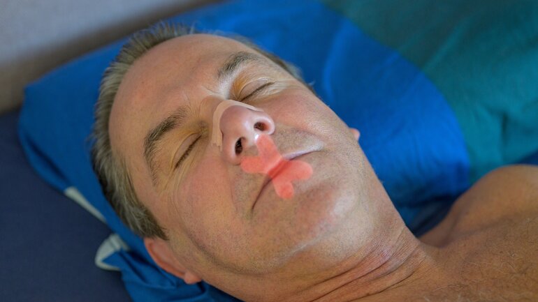 Mann mittleren Alters liegt mit Mouth Tape und Nasenpflaster im Bett und schläft