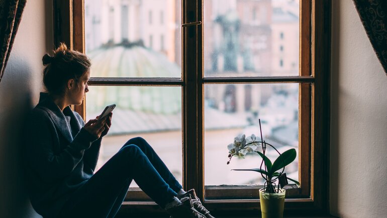 Frau hält Handy in der Hand und schaut aus dem Fenster