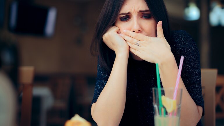 Frau sitzt im Café und hält sich die Hand vor den Mund, da sie nach einem Schluck Cola rülpsen muss