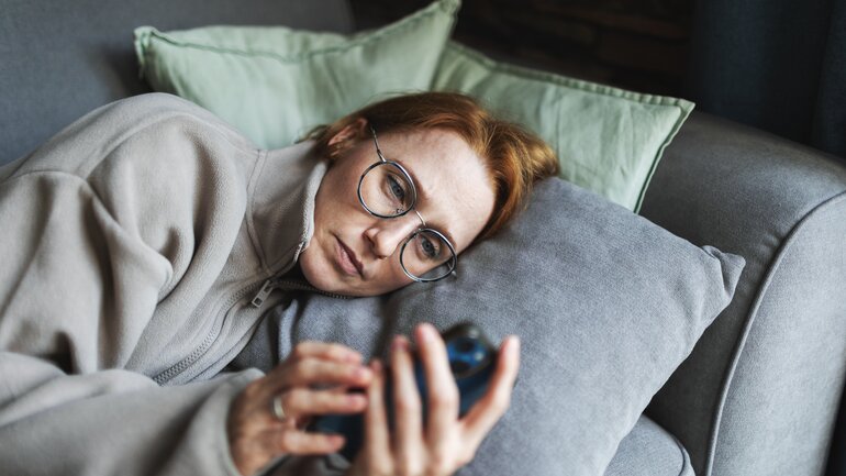 Frau ist sehr müde, liegt auf dem Sofa und schaut auf ihr Smartphone